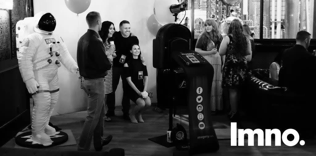 A black and white photo of a photo booth setup with seven people mingling