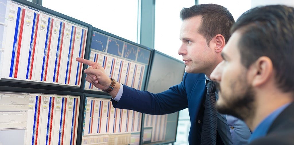 Two millennial white men in suits looking at a grid of monitors 