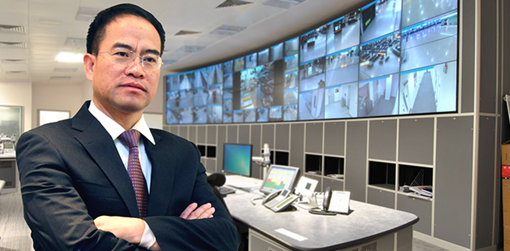An Asian man in a suit in front of a grid of security camera monitors