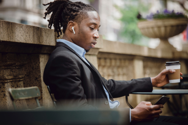 Young black man in suit with headphones on looking at phone and drinking coffee outdoors