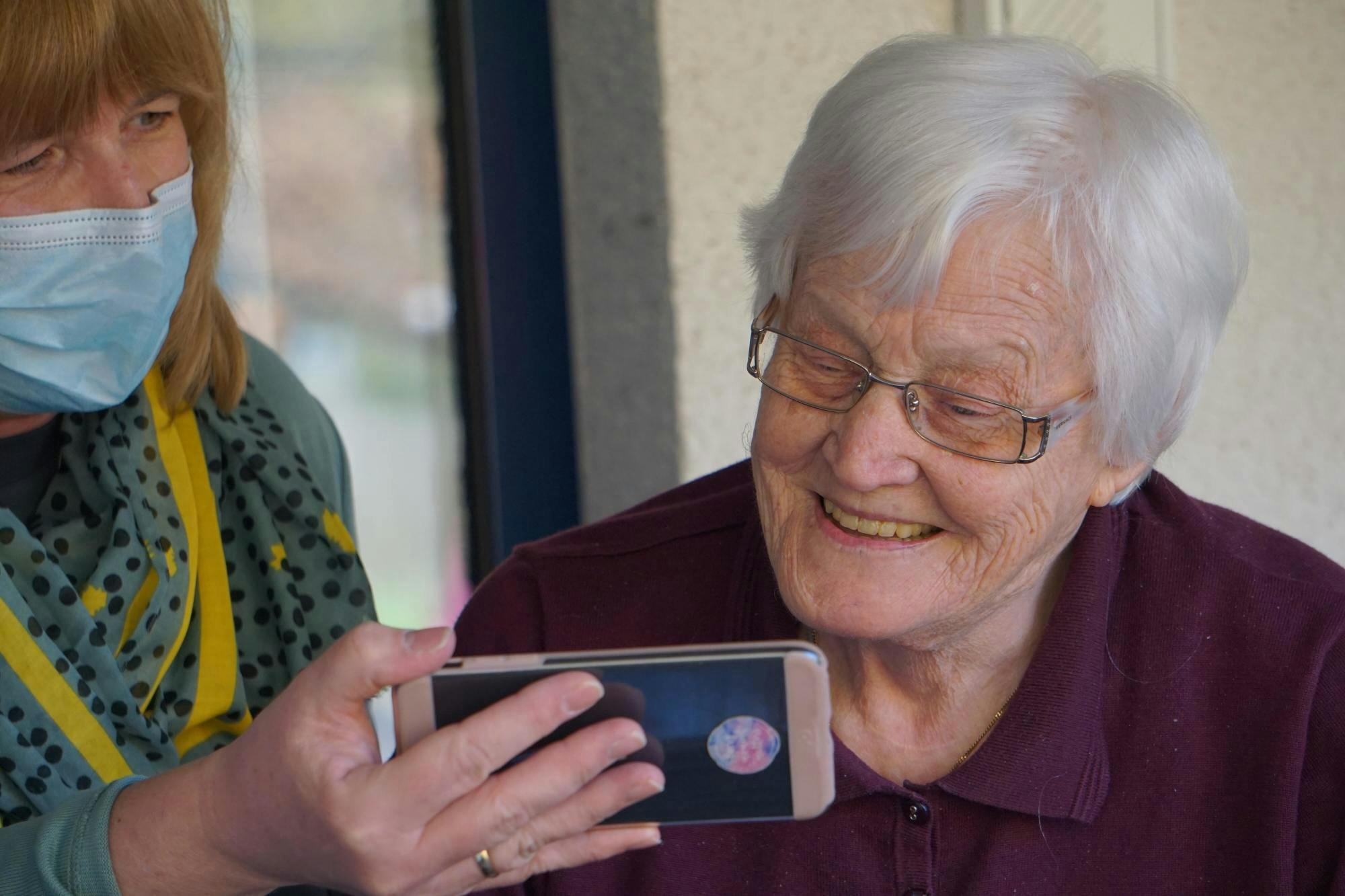 White middle aged woman wearing face mask showing a phone screen to an elderly white woman